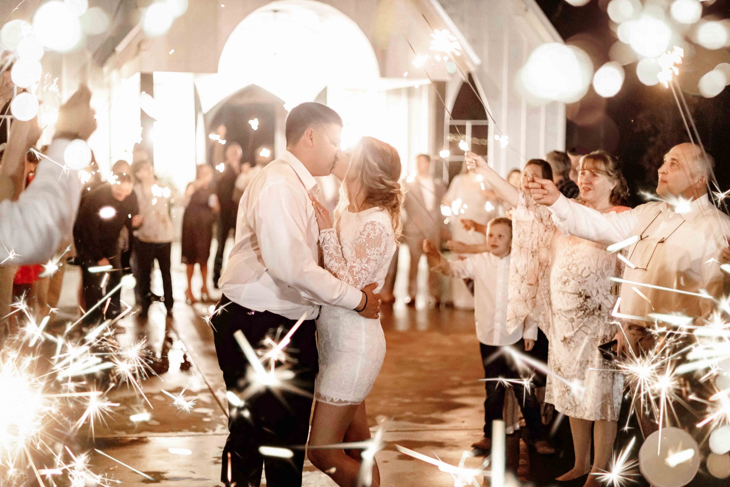 Photo of Bride and Groom kissing as they exit their wedding day among the guests lined up on either side with sparklers shining bright